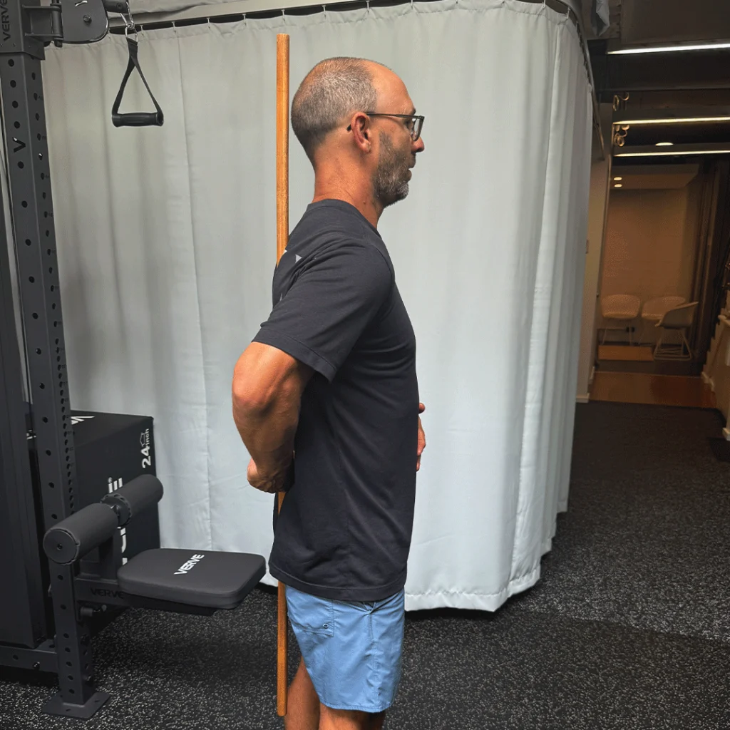 Person in a dark t-shirt and shorts, leaning forward while holding a stick behind their back, performing a hip hinge