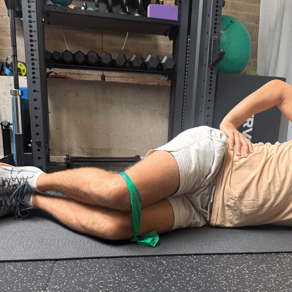 Person lying on their side on a mat, performing a "clam" exercise with a green resistance band around their knees.