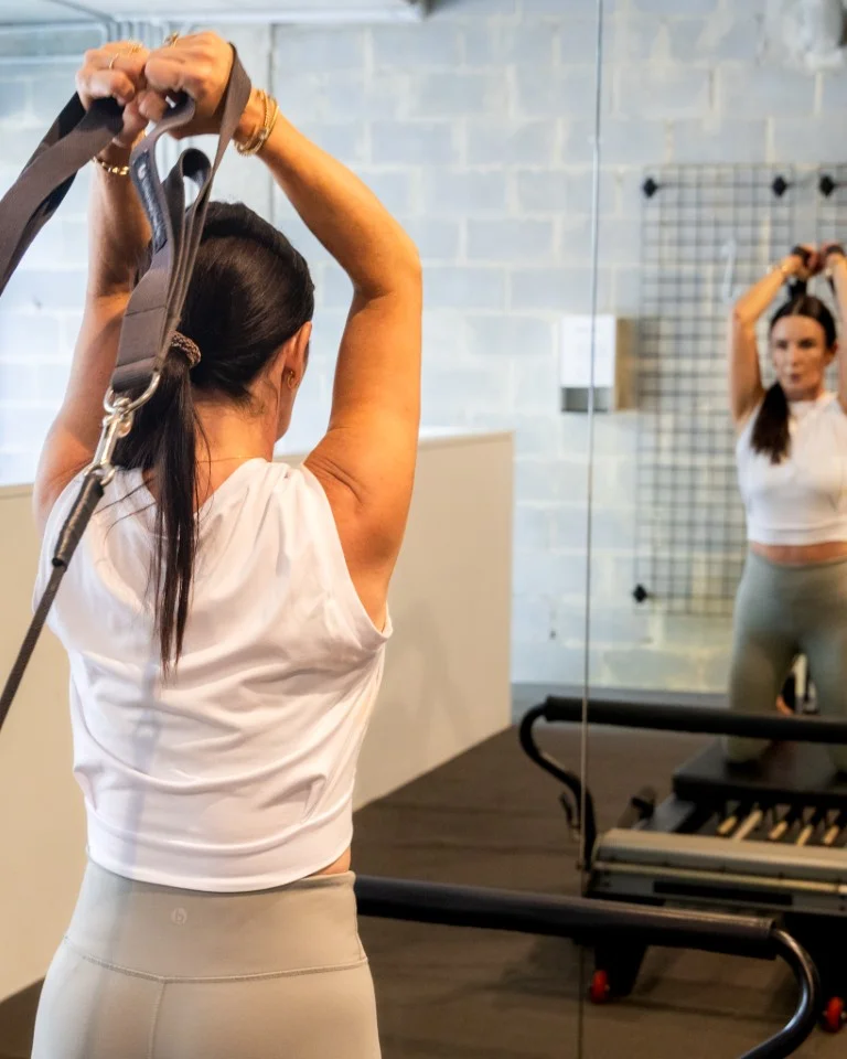 Woman from behind in white activewear, performing an exercise with straps on a Pilates reformer or functional training equipment, with her reflection visible in the mirror