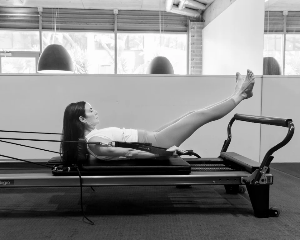 Woman lying supine on a Pilates reformer, with her legs extended upwards, performing an abdominal and leg exercise